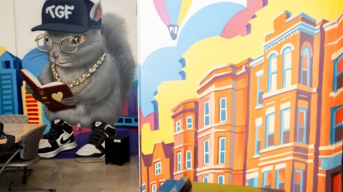 A gray squirrel mascot is shown in a mural wearing a navy cap, gold spectacles, a gold chain and black and white Nikes and reading a red book with a heart on its cover.