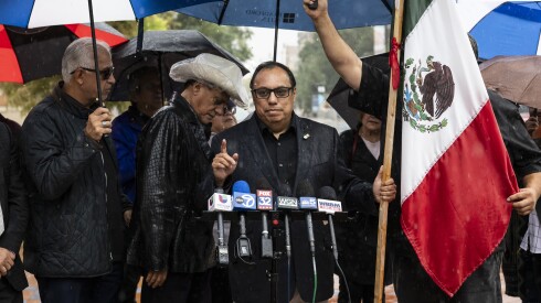 A man wearing glasses speaks into a row of microphones surrounded by other people holding umbrellas and the flag of Mexico.