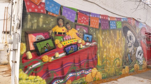 A traditional Día de los Muertos altar, a colorful ofrenda with a red tablecloth, sits in the left corner of the mural, honoring loved ones who have died.