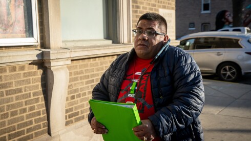 A man with glasses and a blue jacket carries a green binder and talks on his cell phone.