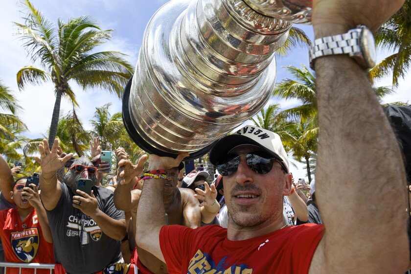 A man in a red T-shirt, baseball hat and glasses is holding a large, tiered silver colored cup. There are people in the background holding out there cellphones to take pictures of the man. Palm trees can also be seen.