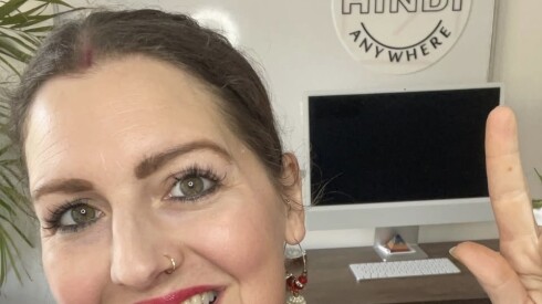 A woman points with both hands to a white circular sign behind her on the wall that reads, "Learn Hindi Anywhere." There is a computer on a desk under the sign.
