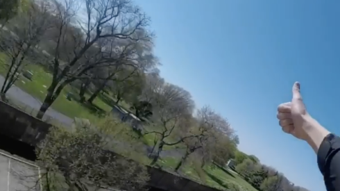 A subway surfer gives a thumbs up from the top of an L car to people on the ground.