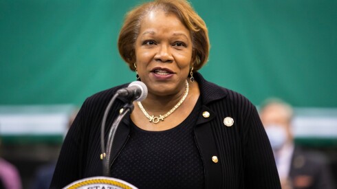 A Black woman with short hair wearing a black cardigan stands in front of a lectern and speaks into a microphone.