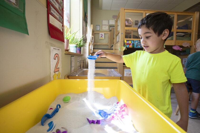 A young boy wearing a yellow T-shirt plays with a toy in a sandbox at an early childhood center.