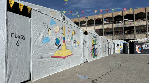 Whites cloths that is the side of tents can be seen. On one of the panels there is a colorful mural. To the left of the mural it reads "class 6." Another building can be seen in the foreground.