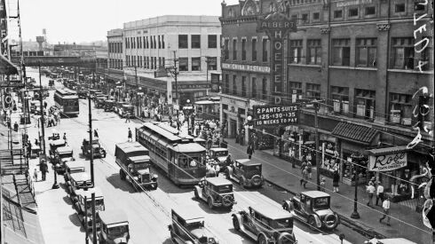 View of automobiles and streetcars driving near the intersection of 63rd and Halsted Streets in the Englewood community area of Chicago, Illinois. Pedestrians are walking on the sidewalks. The Albert Hoefeld store and the Rose Shop are visible on the right side of the image. Text on a sign in front of the Hoefeld store reads: 2 Pants Suits, 25-30-35 dollars, 10 Weeks to Pay.