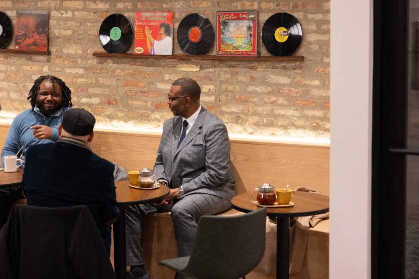 Three Black men sit and talk around a table in a cafe. Vinyl records are displayed on a shelf on the brick wall behind them.