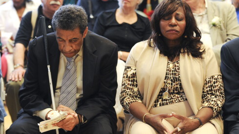 A man and woman are sitting and closing their eyes. The man is in a suit and holding a walking stick. He is looking down. More people can be seen sitting behind the man and woman