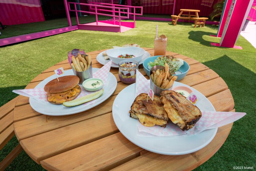 Menu items at the Malibu Barbie Cafe pop-up include the Beach Burger (clockwise from left), the Good Vibes Beet Hummus, the Beach Mojito, the West Coast Wedge Salad, the Live Your Dream Grilled Cheese sandwich and fries, and the Today is the Day Parfait (center).