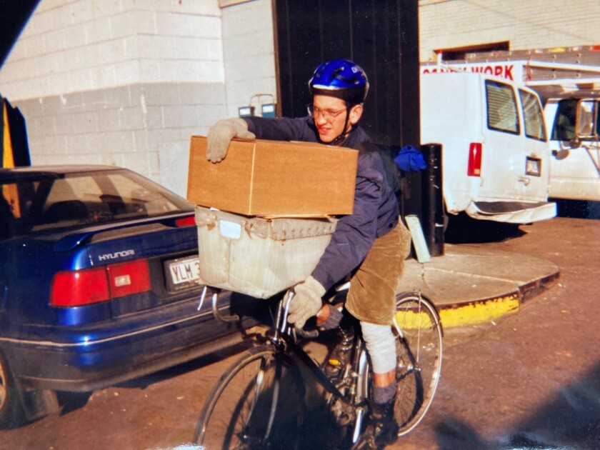 John Greenfield holds a mail bin and cardboard box on the handlebars of his slender bike as he rides through a parking lot.