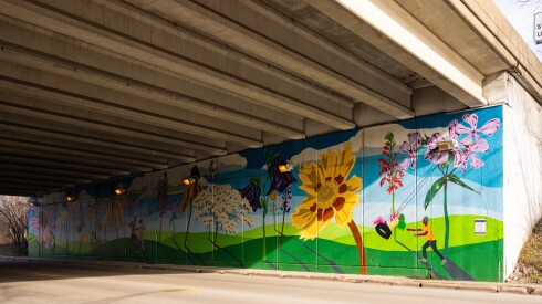 A mural under Interstate 94 depicts a forest of flowers and people doing activities, including a person pushing someone on a tire swing that hangs from one of the flowers.