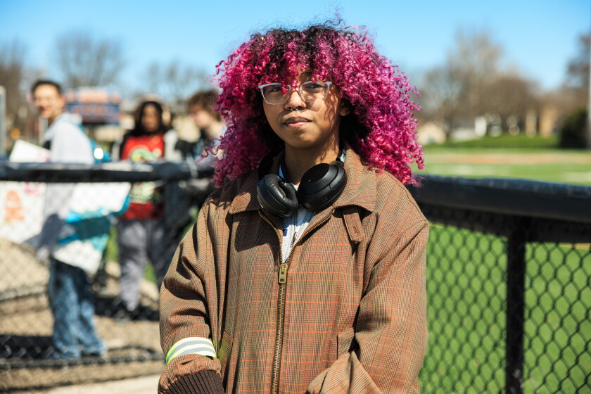 Kevin Watts, a teen wearing headphones and a brown jacket with pink hair, stands near a high school football field.