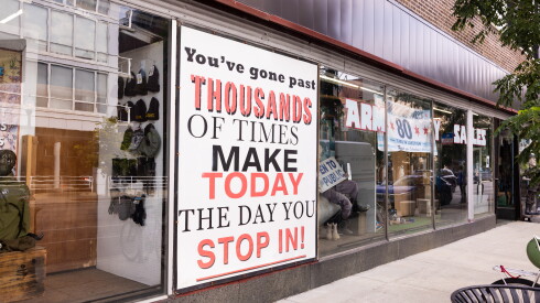Large window sign outside Army Navy Sales store says, "You've gone past thousands of times make today the day you stop in!"