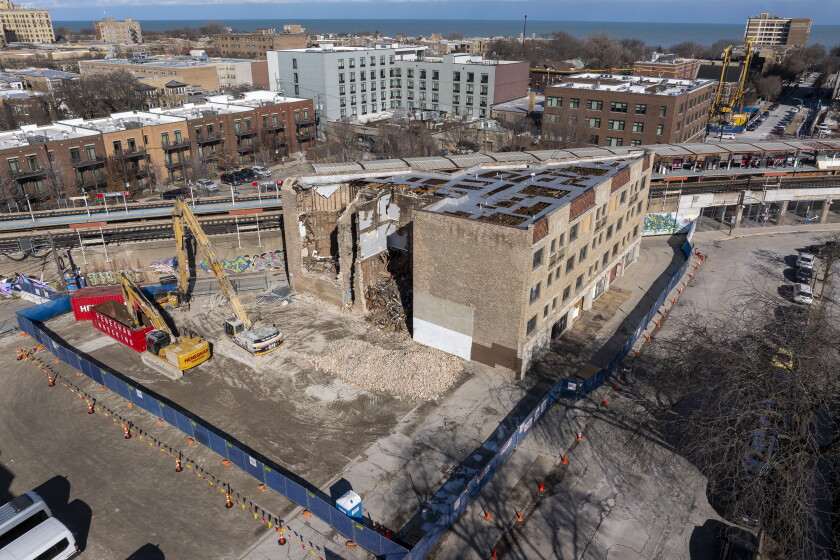 A fence around a partially demolished Rogers Park building with large construction equipment nearby.