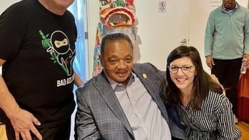 Jesse Jackson, wearing a dress shirt, slacks and blazer, is seated and a woman and man stand at his side.
