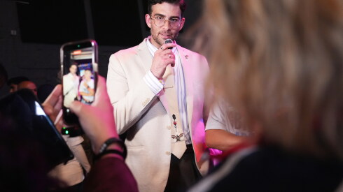 A man in a white suit is speaking into a mic. The back of a person's head is to the right. On the left someone is raising their phone with their hands taking a picture of the man.