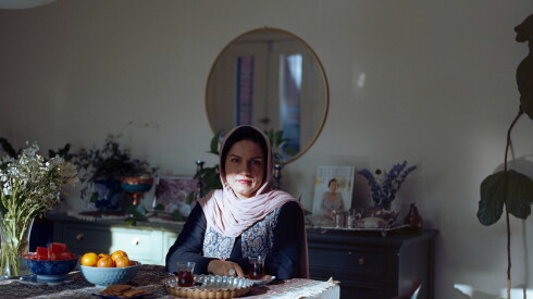A woman in a light colored head covering is sitting at a table covered in a tablecloth. There are bowls of fruits, a tray of biscuit and dark colored tea in a glass on the table.