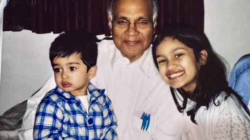 A man is holding a boy in his lap. His arms are clasped around the boy, who is looking away. A little girl, who is smiling, is standing to the right of the man.
