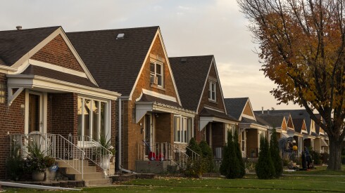 A row of brick homes with green lawns and a tall tree losing its leaves in the fall.