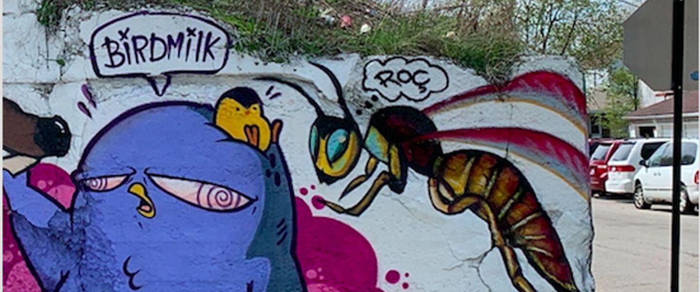 A waving bluebird stands next to a hovering wasp at one end of a Back of the Yards viaduct. The bird design is the signature style of bird_milk_. The wasp was done by an artist who goes by @m157rocio.