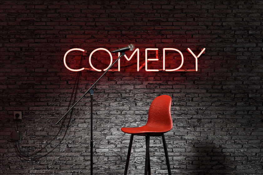 A stage can be seen with a brick lick background. There is red neon sign with the word "COMEDY" on the wall. There is a red chair and microphone in the front.