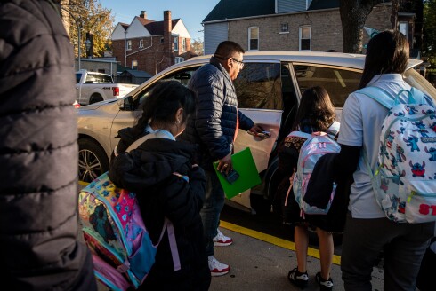 A man wearing a jacket opens the door to a car while four students wearing backpacks wait to get in.