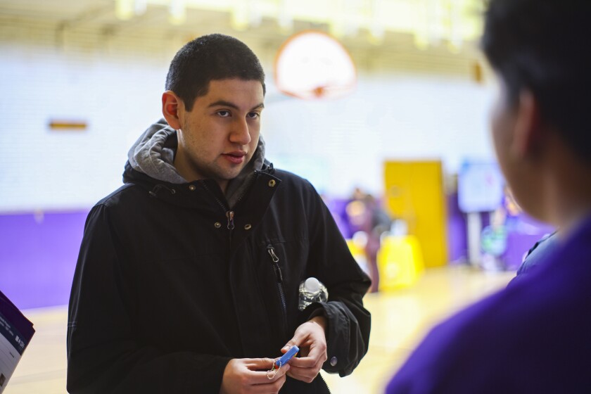 A student wearing a black jacket and holding a keychain speaks with a classmate and stands in a high school gym.