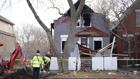 Fire department members work outside a fire-damaged house that’s surrounded by yellow police tape.