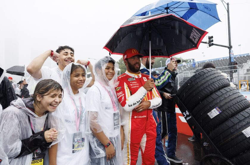 Kids wearing ponchos stand next to NASCAR driver Bubba Wallace in Pit Road posing for a picture.