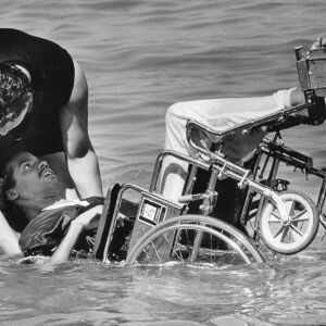 George Anderson, 21, gets an assist from Luis Claudio, 16, as he takes a cool dip, wheelchair and all, at Oak Street Beech in July of 1986.