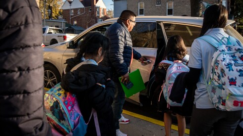 A man wearing a jacket opens the door to a car while four students wearing backpacks wait to get in.