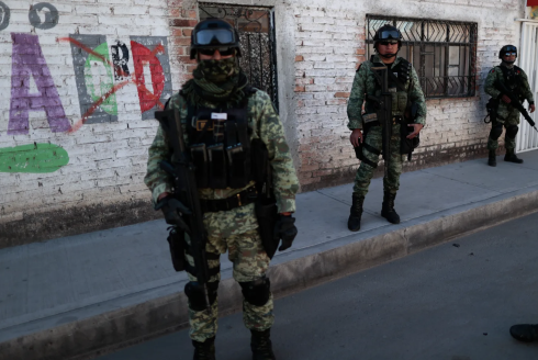 Soldiers wearing green camouflage and tactical equipment stand on the sidewalk and road in front of a white brick building Irapuato, Mexico.