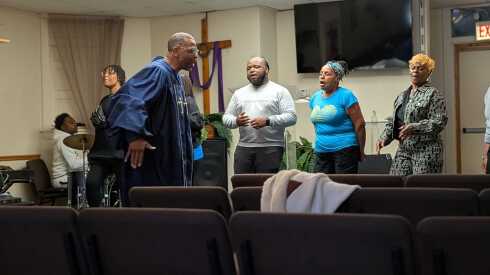 A Black pastor wearing blue robes sings in a storefront church backed by singers and a drummer.