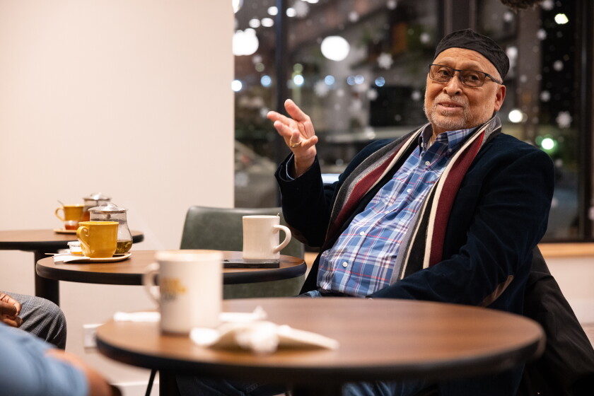A light-skinned Black mean wearing a blue checkered shirt, a scarf and a blazer gestures with his hand while seated at a table topped with a coffee cup.