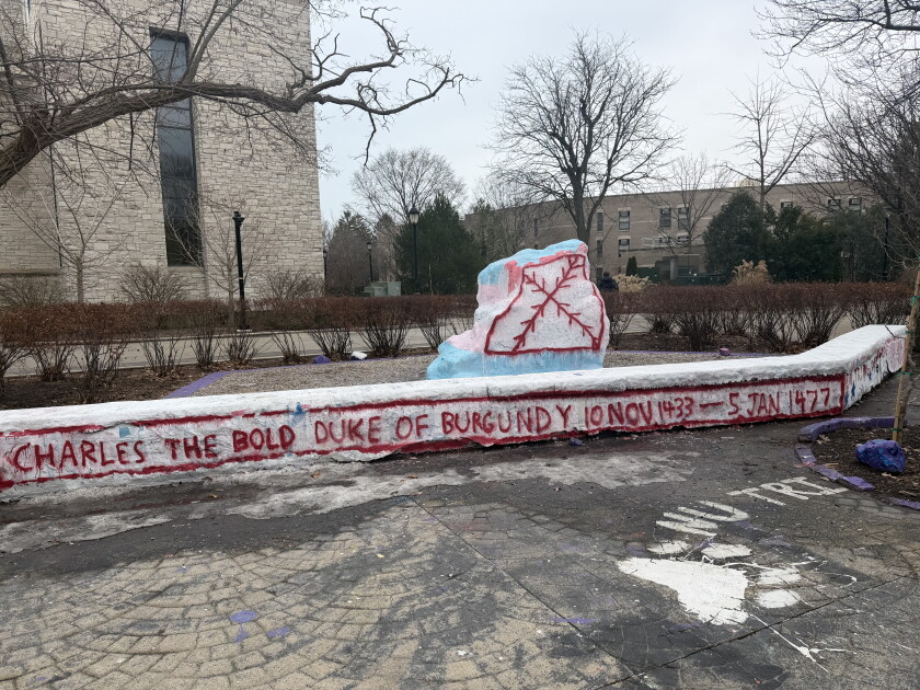 A giant boulder on a college campus is painted over with a red jagged cross while bricks nearby bear the words "Duke of Burgundy."