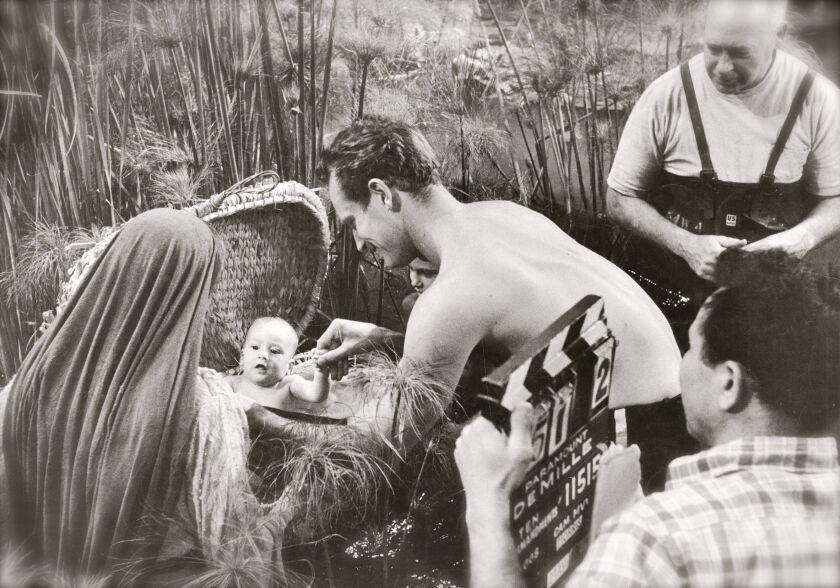 As director Cecil B. DeMille (background) looks on, the baby Moses, played by Fraser Heston, is tended to by his famous real-life dad Charlton Heston during this scene being filmed on the backlot of Paramount Studios in 1955.