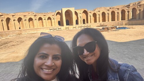Two woman are smiling in a selfie in front of sand-colored ancient ruins. The woman on the right has sunglasses on. The woman on the left is wearing them on her head.