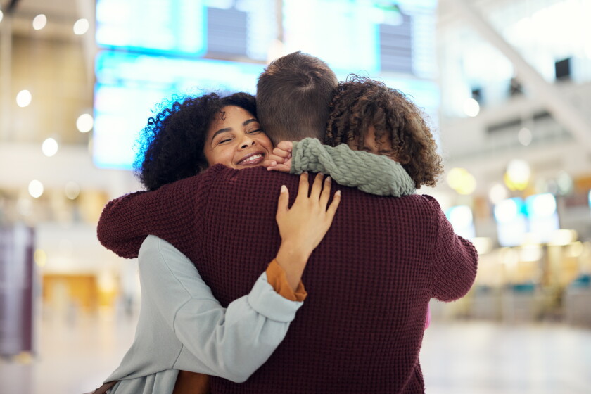The back of a man embracing a woman and child. The woman's face can be seen. Her arm in on the man's back. The child's face can't be seen as it is is nestled in the man's neck.