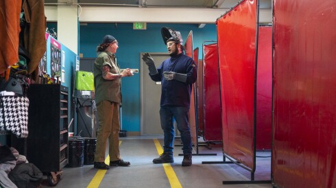 A trainee talks to an instructor wearing a welding mask in a factory.