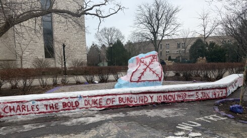A giant boulder on a college campus is painted over with a red jagged cross while bricks nearby bear the words "Duke of Burgundy."