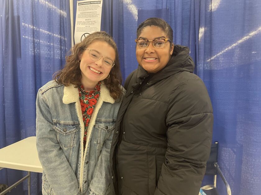 Two women, Meredith Metzger and Mari Oliver, smile in front of a blue curtain at a voting center.