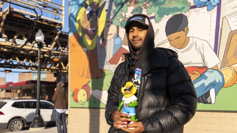 Muralist Kari Blak stands next to his painting on the Bessie Coleman Branch Library, holding a 3D print of his character named Taru which is the mascot of his company Petalhead.