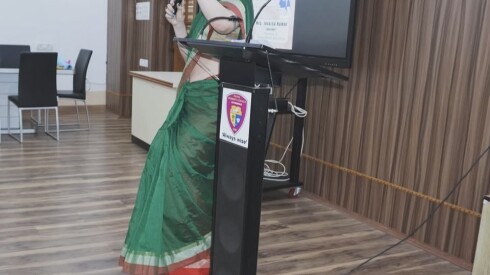 A white woman in a green sari speaks near a podium with a thin mic. There is a screen behind the woman.