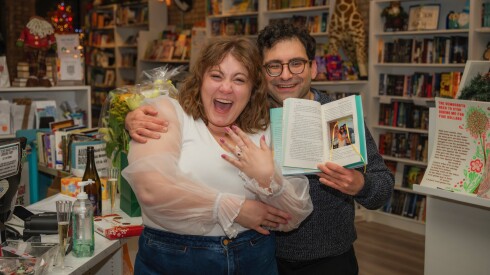 A woman shows her engagement ring next to a man holding an open book in a bookstore.