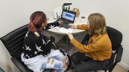 A teen wearing a black shirt with white stars and ripped jeans sits at the computer with an adult woman wearing a mustard jacket and black pants.