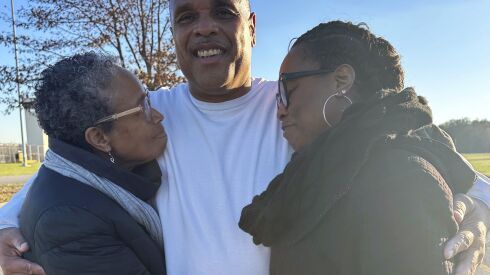 Brian Beals, wearing a white long-sleeved shirt, hugs his sister Pattilyn and his niece Tamiko, clad in coats, outside Robinson Correctional Institution, with blue skies and trees in the background on a sunny day.