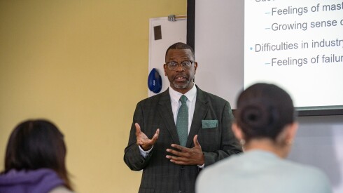 A Black man wearing glasses and a suit and tie gestures with his hands while teaching a college class.