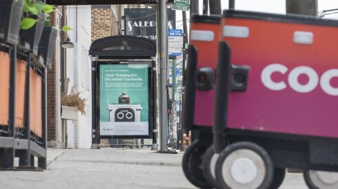 A pair of Coco Delivery Robots are parked Tuesday near a new advertisement on the eastern side of a CTA bus shelter at West Grand Avenue and North Racine Avenue. The ad displays an apology from competing delivery robot company Serve Robotics for one of its delivery robots damaging the bus shelter last month.