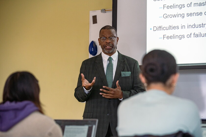 A Black man wearing glasses and a suit and tie gestures with his hands while teaching a college class.